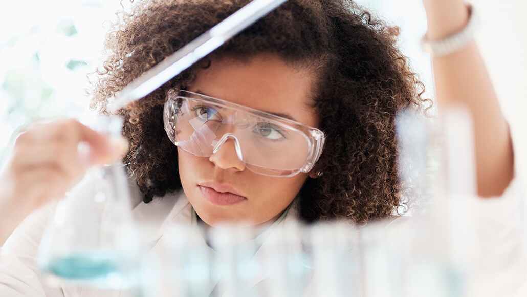 A female scientist with goggles in her lab examining substances in beakers.